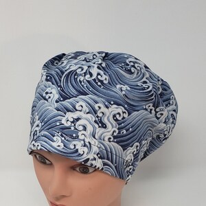 May include: A blue and white wave-patterned scrub cap. The cap is made of fabric and covers the head. The design features a repeating pattern of stylized ocean waves in shades of blue and white.