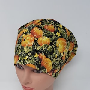 May include: A patterned scrub cap featuring a repeating design of orange pumpkins, green leaves, and yellow flowers on a black background. The cap is worn on a mannequin head.