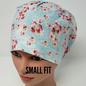 May include: A light blue scrub cap featuring a floral pattern with pink and yellow flowers. The cap is displayed on a mannequin head, with the text "SMALL FIT" in black.