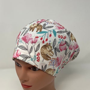 May include: A white surgical scrub cap with a floral and sloth pattern. The cap features pink flowers, green leaves, and brown sloths. The cap is designed to cover the head and hair.