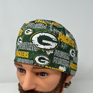 May include: A scrub cap featuring a green background with a repeating pattern of the white and yellow Green Bay Packers logo and the word PACKERS in white and yellow text. The fabric has a distressed, textured appearance throughout the design.