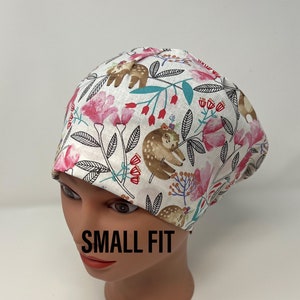 May include: A white scrub cap with a floral and sloth pattern. The cap features pink flowers, green leaves, and brown sloths. The text "SMALL FIT" is printed on the front of the cap.