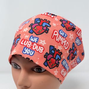 May include: A coral-colored surgical scrub cap with a repeating pattern of stylized hearts and text. The text includes phrases like "We Lub-Dub You" and "Get Pumped." The cap is designed for medical professionals.