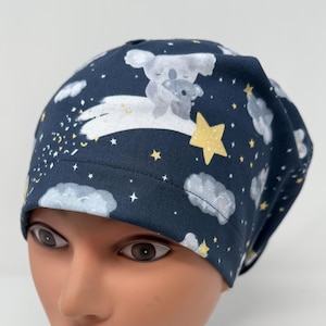 May include: A navy blue scrub cap featuring a whimsical design of koalas, stars, and clouds. The koalas are depicted in a soft gray, with yellow stars and white clouds scattered across the fabric. The cap is designed to fit comfortably.