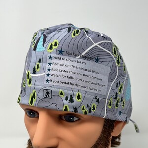 May include: A gray and blue patterned surgical scrub hat with a white label that reads "Trail Park Etiquette" and lists rules for trail use. The rules include "Yield to slower bikers", "Remain on the trails at all times", "Ride faster than the bears can run", "Watch for fallen rocks and avoid them", and "If you pedal harder you'll speed up".