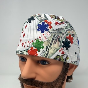 May include: A white surgical scrub cap with a repeating pattern of playing cards, poker chips, and dollar bills. The cap features red, green, blue, and black poker chips, and various playing card suits.