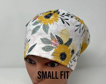 May include: A white scrub cap with a yellow sunflower pattern, green leaves, and pink accents. The cap is on a mannequin head. The text "SMALL FIT" is printed on the mannequin.