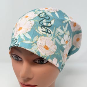 May include: A teal blue scrub hat with a white floral pattern and the name "BRIA" embroidered in black.