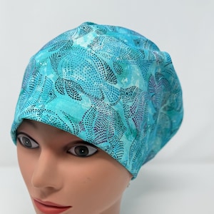 May include: A turquoise and teal patterned scrub cap featuring a sea turtle design. The cap is made of a soft fabric and is designed to fit comfortably. The cap is shown on a mannequin head.