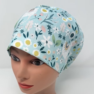 May include: A light blue surgical scrub cap with a floral and llama pattern. The cap features white, pink, and yellow flowers, green leaves, and white llamas. The cap is designed to cover the head and hair.