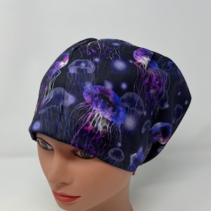 May include: A black surgical scrub cap featuring a vibrant pattern of purple and blue jellyfish. The jellyfish have long, flowing tentacles and are set against a dark background with glowing orbs. The cap is designed to fit snugly on the head.
