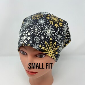 May include: A black cap featuring a snowflake design in gold and white. The cap is displayed on a mannequin head. The text "SMALL FIT" is printed on the mannequin's face.