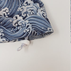 May include: A blue and white patterned fabric item, possibly a hat or accessory, featuring a wave design. The fabric has a drawstring with a white toggle. The item is set against a white background.