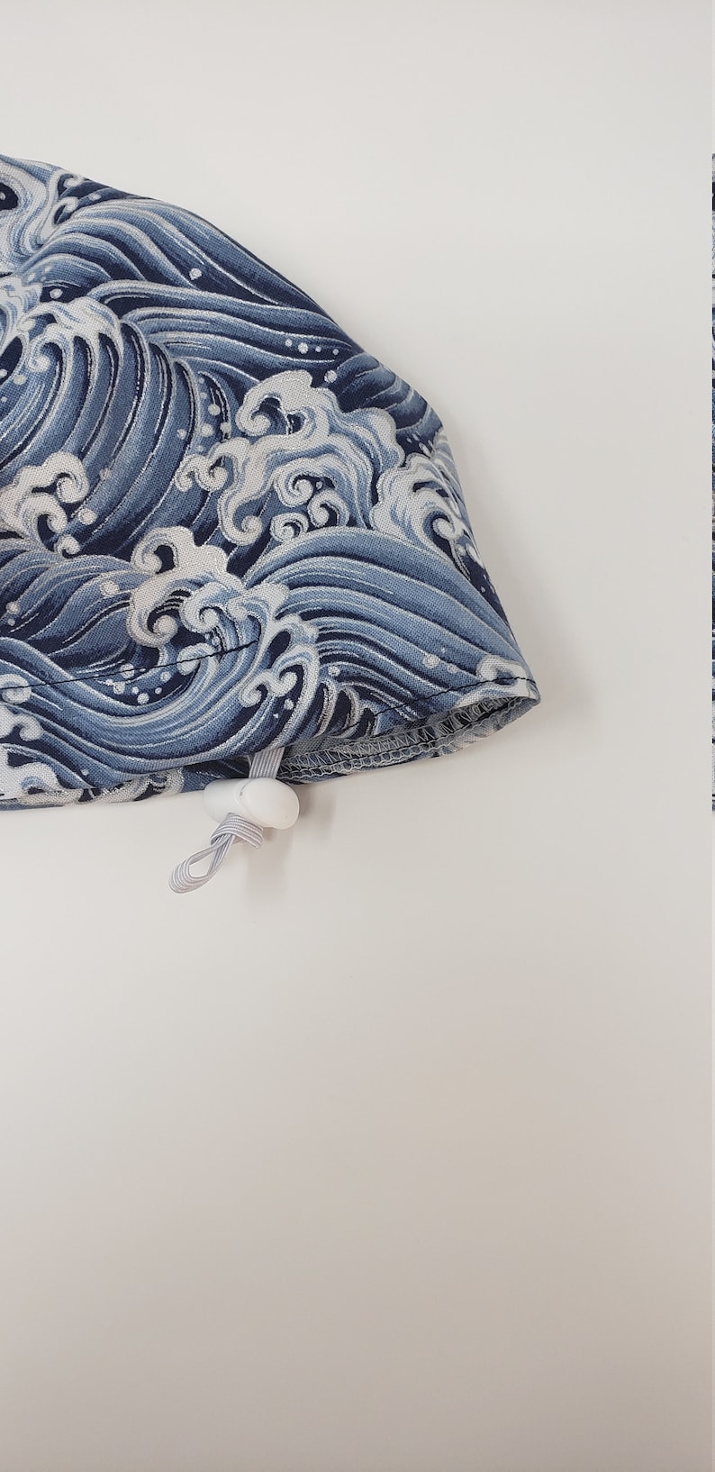 May include: A blue and white patterned fabric item, possibly a hat or accessory, featuring a wave design. The fabric has a drawstring with a white toggle. The item is set against a white background.