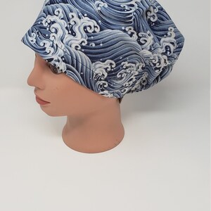 May include: A blue and white wave-patterned hat is displayed on a mannequin head. The hat features a repeating design of stylized ocean waves in shades of blue and white. The hat is a rounded shape with a small brim.