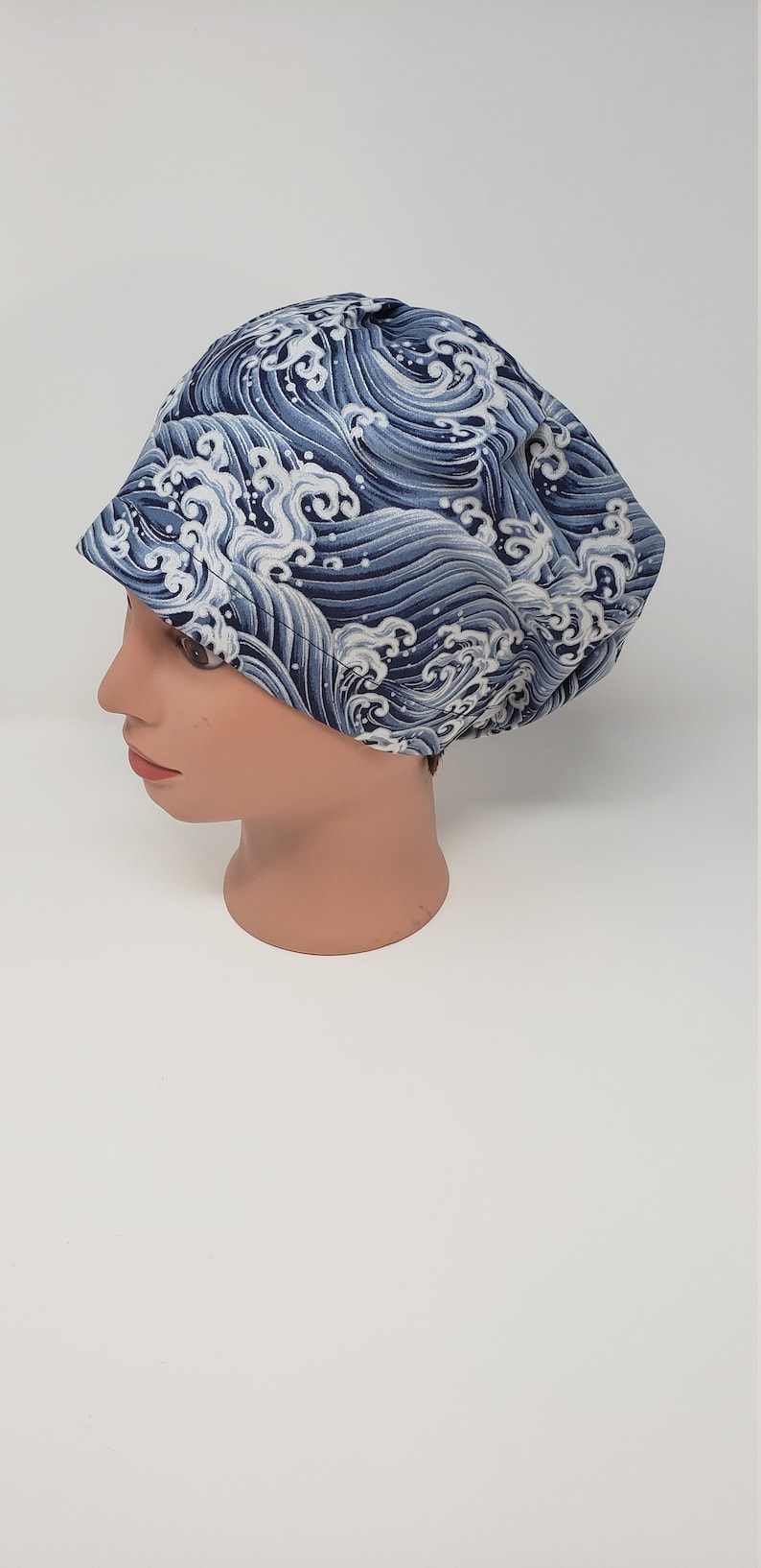 May include: A blue and white wave-patterned hat is displayed on a mannequin head. The hat features a repeating design of stylized ocean waves in shades of blue and white. The hat is a rounded shape with a small brim.