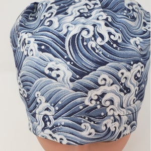 May include: A blue and white wave-patterned scrub cap. The fabric design features stylized ocean waves in shades of blue and white. The cap is designed to fit snugly on the head, with a rounded shape.