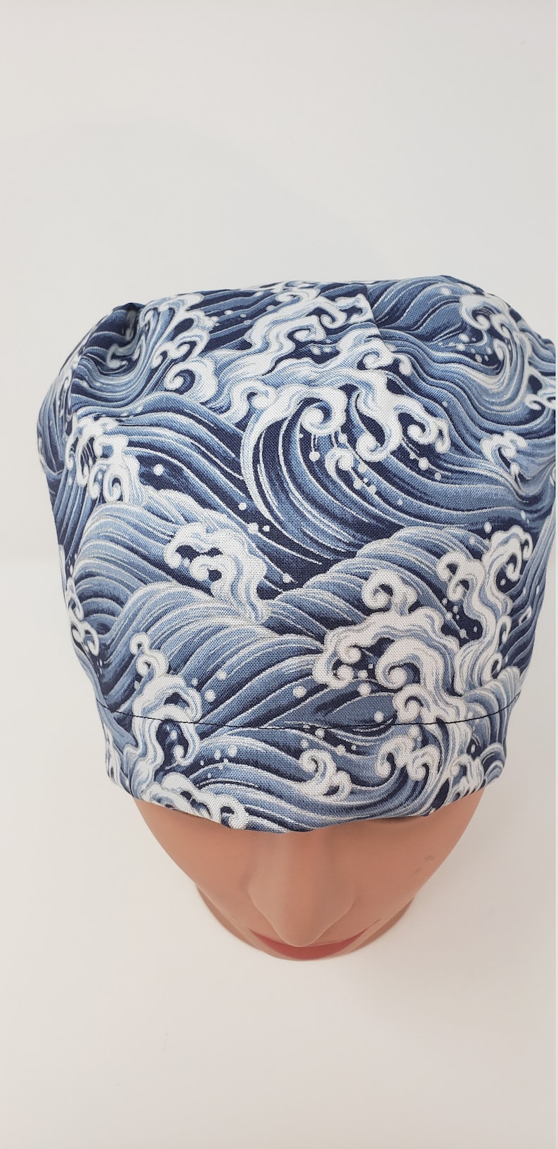 May include: A blue and white wave-patterned scrub cap. The fabric design features stylized ocean waves in shades of blue and white. The cap is designed to fit snugly on the head, with a rounded shape.