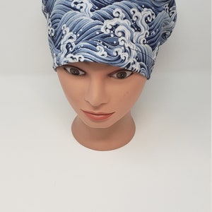 May include: A blue and white patterned scrub cap featuring a wave design. The cap is made of fabric and is displayed on a mannequin head. The wave pattern is a repeating design in shades of blue and white.