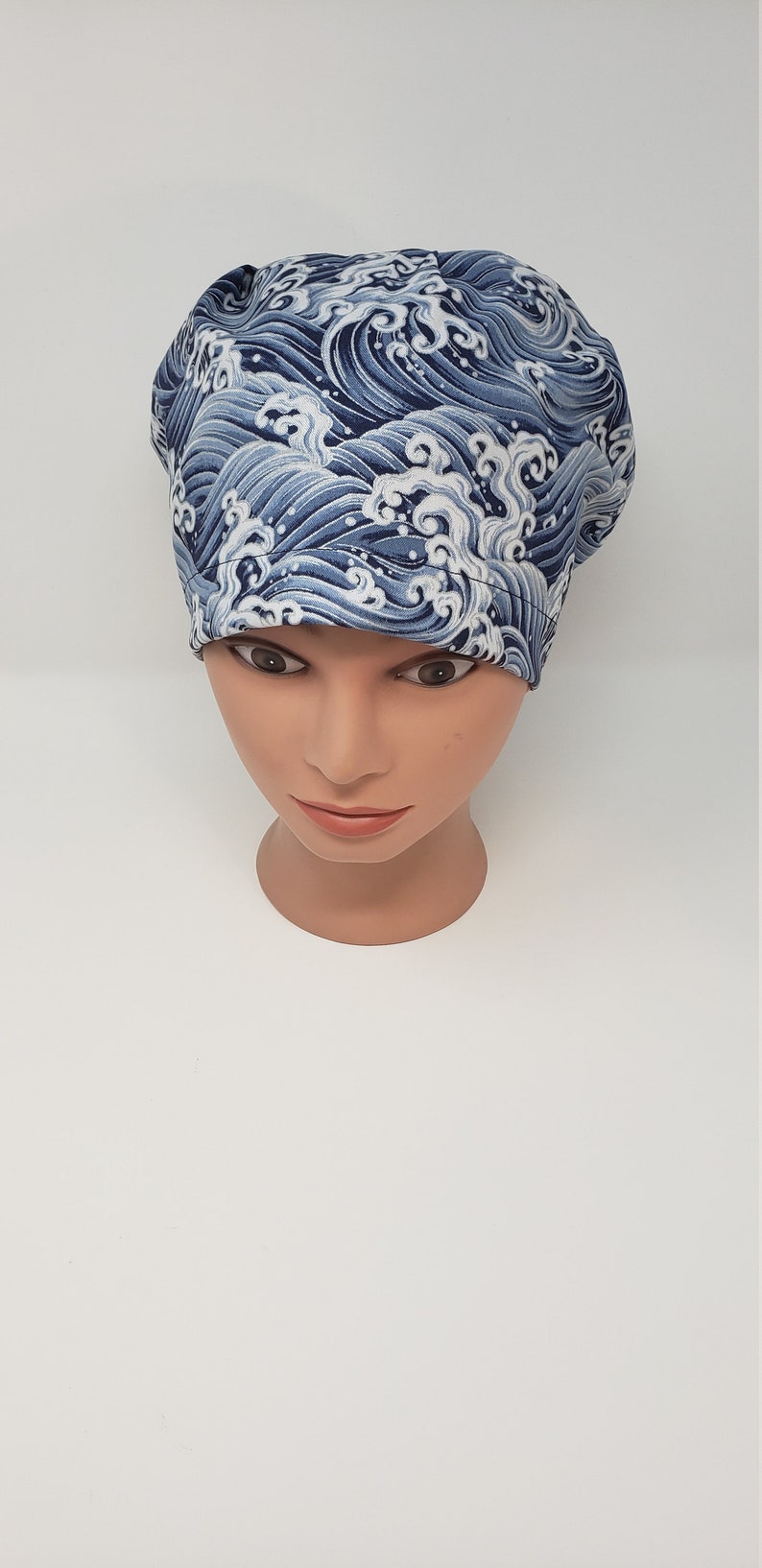 May include: A blue and white patterned scrub cap featuring a wave design. The cap is made of fabric and is displayed on a mannequin head. The wave pattern is a repeating design in shades of blue and white.