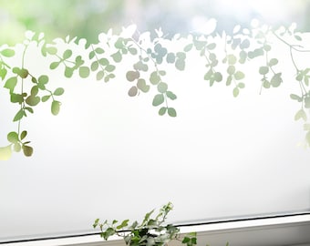 May include: A window with a frosted film featuring a green leaf design. A small potted plant in a woven basket and a decorative candle holder sit on the windowsill. The background is blurred, suggesting an outdoor scene.