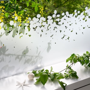 Op de afbeelding: Een wit gematteerd raamfolie met een bladmotief. De folie is aangebracht op een raam met uitzicht op groen gebladerte buiten. Een kleine tak met groene bladeren en een wit decoratief voorwerp liggen op een wit oppervlak onder het raam.