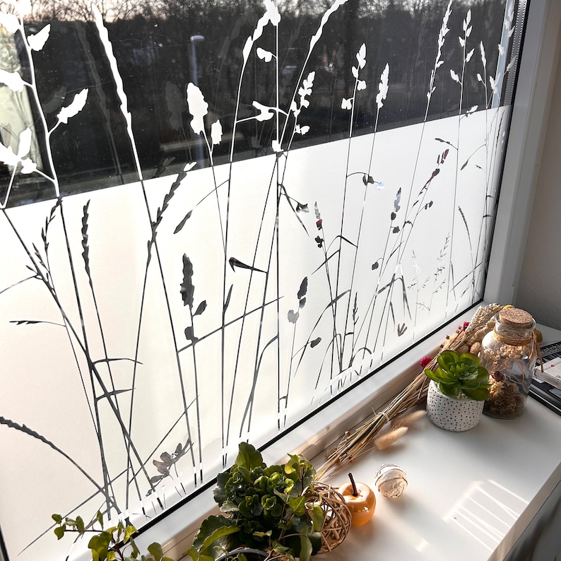 May include: A white window with a frosted glass panel featuring a silhouette of tall grass. The windowsill is decorated with dried flowers, a small potted plant, and a glass jar.