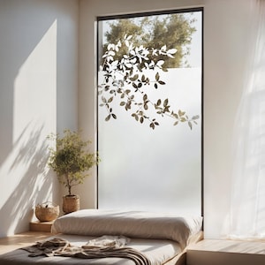 Könnte beinhalten: Ein weißes, mattes Fenster mit einer Silhouette eines belaubten Astes. Im Vordergrund ein Bett mit einer weißen Bettwäsche und einer braunen Wolldecke.
