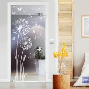 Op de afbeelding: Een matglazen deur met een wit silhouetontwerp van paardenbloemen en vlinders. De deur is wit omlijst en staat tegen een muur met een houten lattenwand en een witte vaas met gele bloemen.