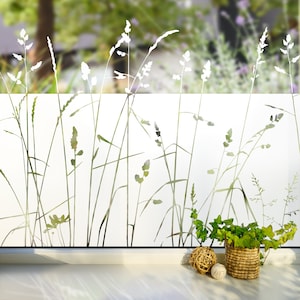 May include: White frosted window film with a silhouette design of tall grass and wildflowers. The film is installed on a window with a white windowsill and a potted plant in front of it.