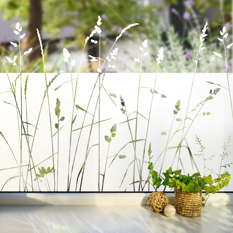 May include: White frosted window film with a silhouette design of tall grass and wildflowers. The film is installed on a window with a white windowsill and a potted plant in front of it.