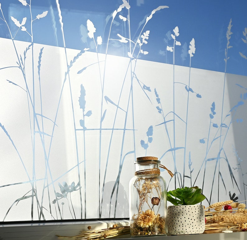 May include: A clear glass jar with a cork lid filled with dried flowers and plants sits on a windowsill. The window has a frosted glass film with a design of tall grass silhouettes.