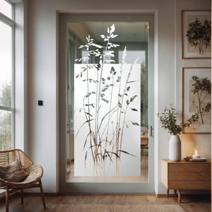 Peut inclure: Une porte en verre givré avec une silhouette blanche de hautes herbes et de fleurs. La porte est dans une maison moderne avec un sol en bois et une table d'appoint en bois.