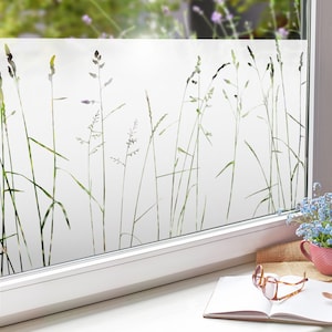 Könnte beinhalten: Eine Milchglasfolie mit einem Silhouetten-Design aus hohem Gras in Grüntönen. Die Folie ist auf ein Fenster mit weißem Rahmen angebracht. Ein Buch, eine Brille und ein kleiner Strauß blauer Blumen in einer rosa Vase stehen auf der Fensterbank.