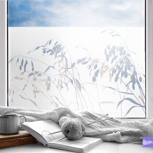Könnte beinhalten: Ein Fenster mit einem mattierten Glasdesign, das eine Silhouette von hohem Gras mit blauen und braunen Akzenten zeigt. Eine weiße Decke, ein Buch und eine Wollknäuel liegen auf einer Fensterbank vor dem Fenster.