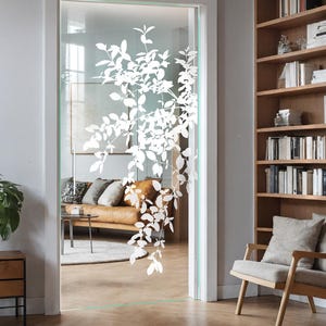May include: A frosted glass door with a white floral design etched into the glass. The door is open and shows a living room with a brown leather sofa and a white rug. A wooden chair with a light grey cushion sits in front of a wooden bookshelf filled with books.