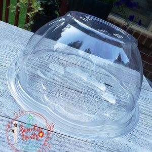 May include: A clear, plastic dome-shaped container with a scalloped base. The container is transparent, allowing a view of the contents. The base has a slightly raised edge. The container is designed for food storage or display.