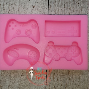 May include: Pink silicone mold with four different video game controller designs. The mold includes a classic NES controller, a Playstation controller, an Xbox controller, and a retro game console.