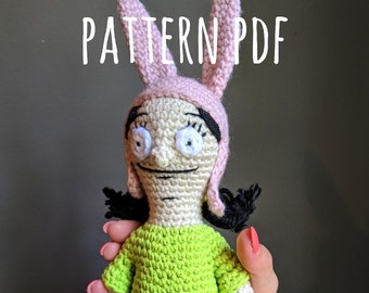 May include: Crocheted amigurumi doll with pink bunny ears, a green dress, and black shoes. The doll has a white face with black eyes and a black mouth. The doll is being held by a hand with pink nail polish.