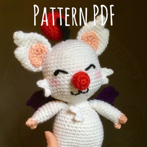 May include: White crocheted amigurumi toy with a red nose, red hat, and purple wings. The toy is being held in a hand.