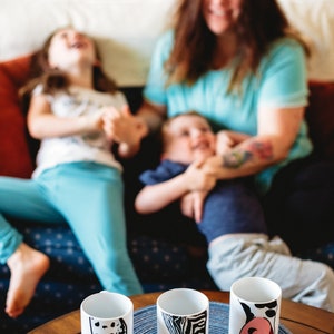 May include: Three white ceramic mugs with different animal designs. The mugs are on a wooden table with a blue and white placemat. The mugs are decorated with a black and white dalmatian, a black and white zebra, and a black and white cow.