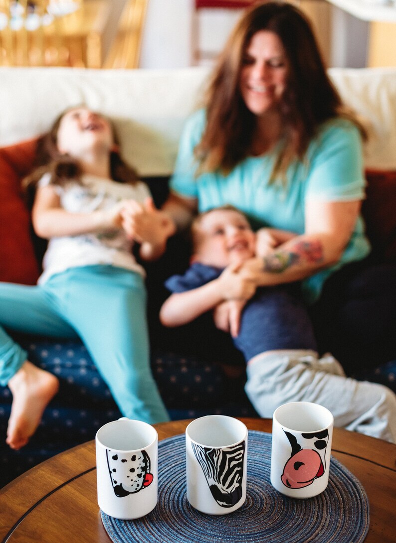 May include: Three white ceramic mugs with different animal designs. The mugs are on a wooden table with a blue and white placemat. The mugs are decorated with a black and white dalmatian, a black and white zebra, and a black and white cow.