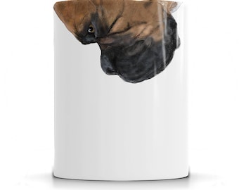 May include: White ceramic mug with a brown and black dog's head printed on it. The dog's head is upside down and the dog is looking at the camera.