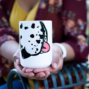 May include: A white ceramic mug with a black and white dalmatian dog face design. The dog's tongue is pink and sticking out. The mug is a standard size and has a rounded handle.
