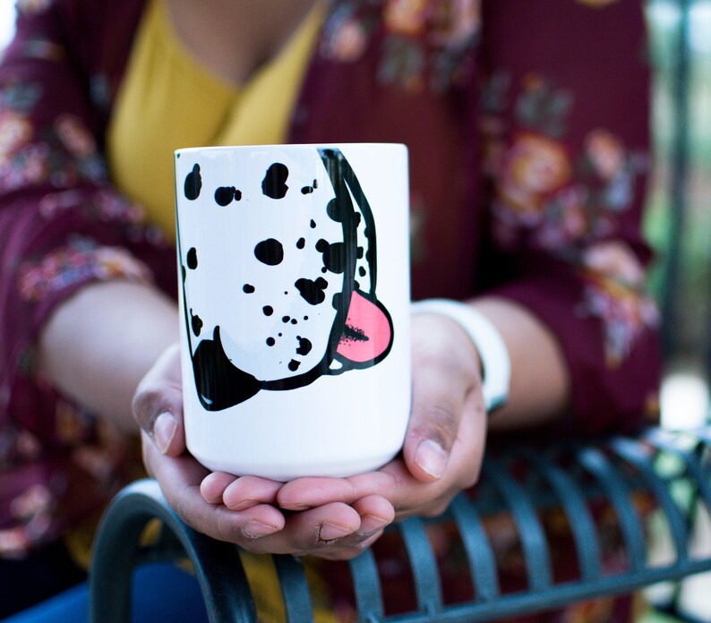 May include: A white ceramic mug with a black and white dalmatian dog face design. The dog's tongue is pink and sticking out. The mug is a standard size and has a rounded handle.