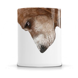 May include: A white ceramic mug with a brown and white dog's face peeking out from the side. The dog's face is looking down at the mug.