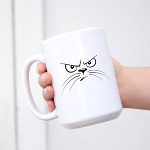 May include: White ceramic mug with a black and white illustration of an angry cat face.