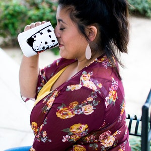 May include: A white ceramic mug with a black and white dalmatian dog design. The mug is filled with a beverage and the person is drinking from the mug.