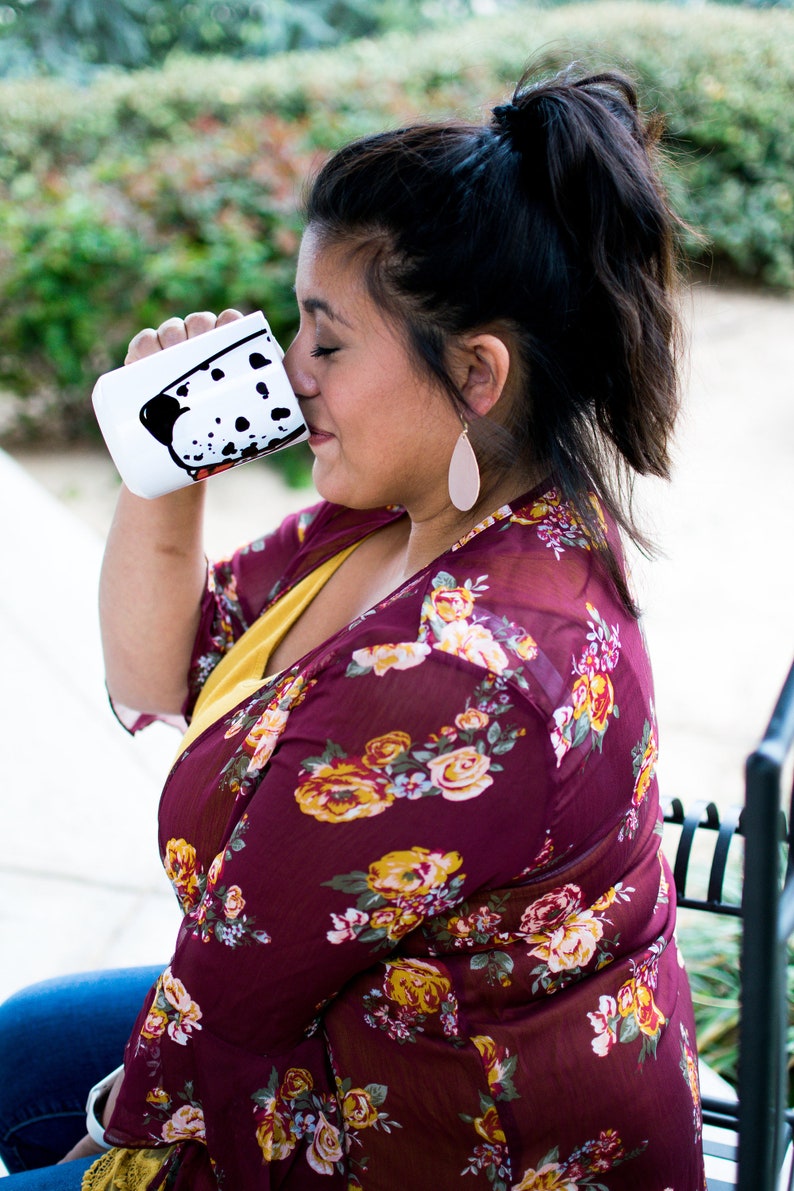 May include: A white ceramic mug with a black and white dalmatian dog design. The mug is filled with a beverage and the person is drinking from the mug.