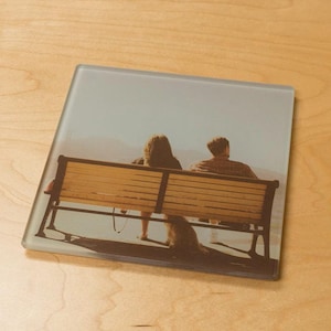 May include: Square glass coaster featuring a photograph of two people sitting on a park bench, with a dog at their feet. The bench is made of wood and metal, and the background shows a mountain range and a light blue sky.